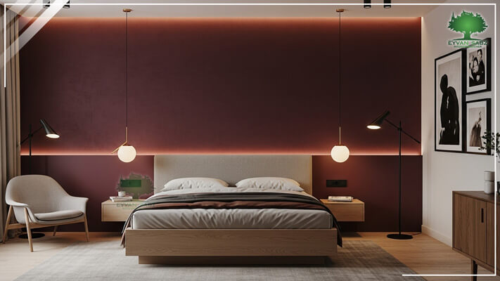 اتاق خواب مینیمال با دیوار تاکیدی (Accent Wall) به رنگ زرشکی تیره و نورپردازی خطی مخفی. تخت خواب با قاب چوبی روشن، چراغهای آویز کروی و صندلی مدرن.